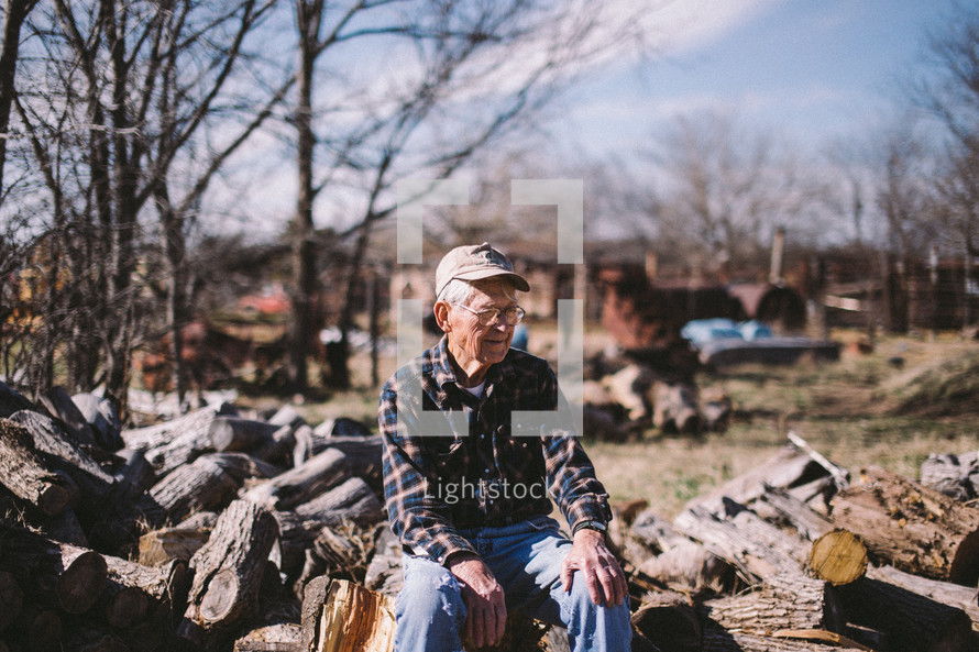 an elderly man sitting on a pile of cut logs 