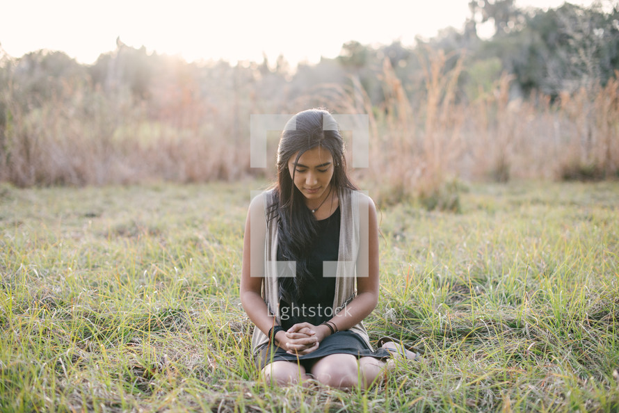 A young woman sitting and praying in a field of grass.