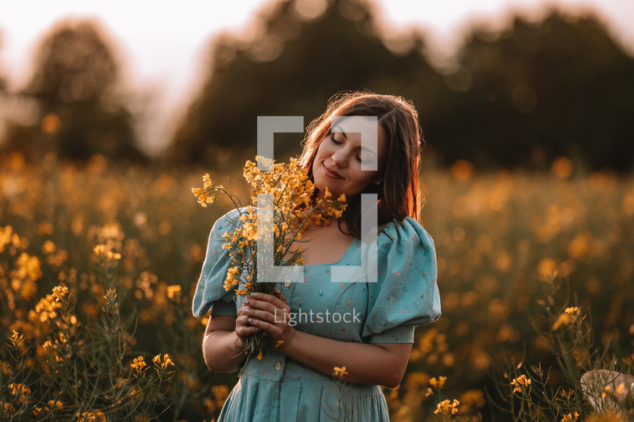 Portrait of attractive woman posing in blooming canola flowers field. Elegant girl in retro dress with bouquet, countryside nature place. Rapeseed meadow, vintage outfit, spring season