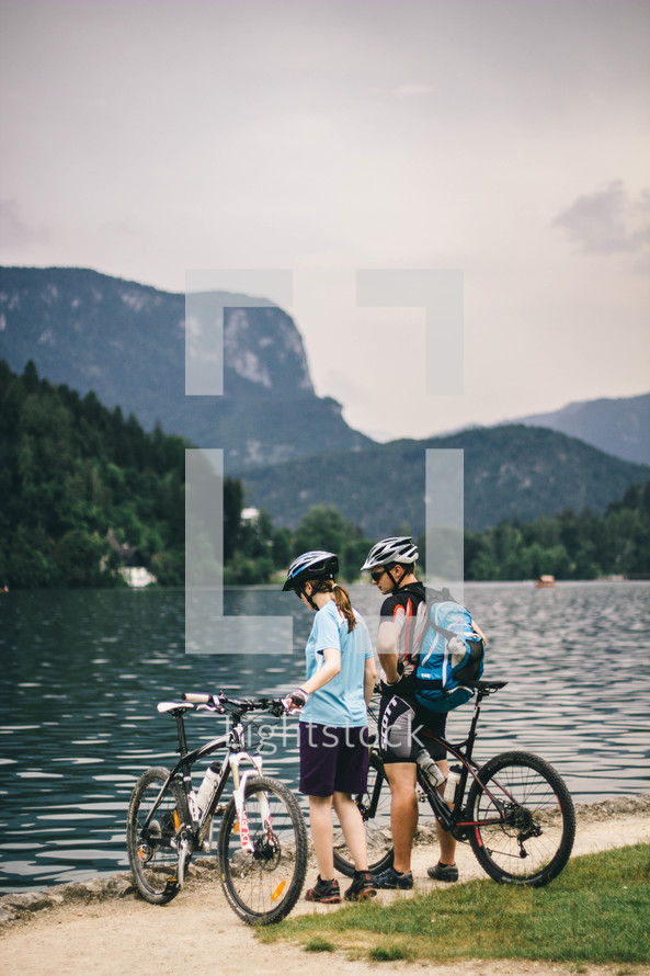 Two people with bicycles by a lake in the mountains.
