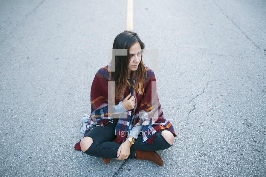 a teen sitting in the middle of a road wrapped in a blanket 
