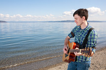 Smiling man playing guitar at water's edge.