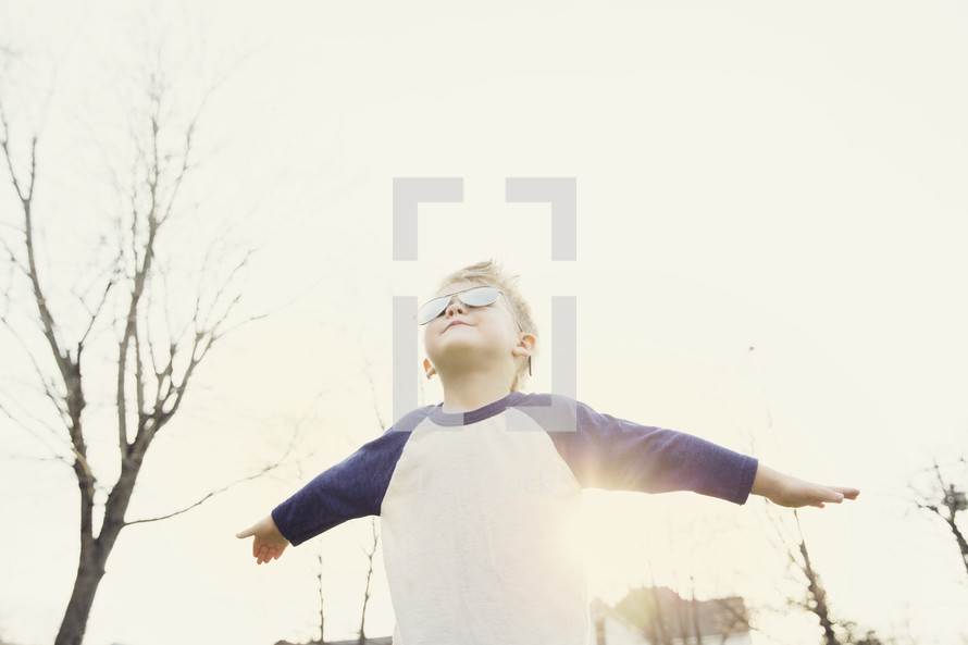 A little boy in sunglasses stands in the sun with arms outstretched.