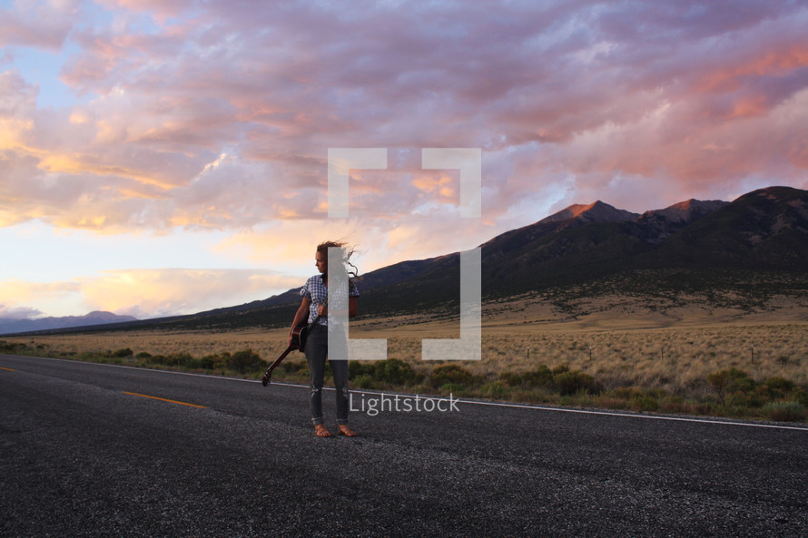 a young woman standing in the middle of a road holding a guitar 