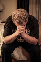 Man praying on the edge of a bed.