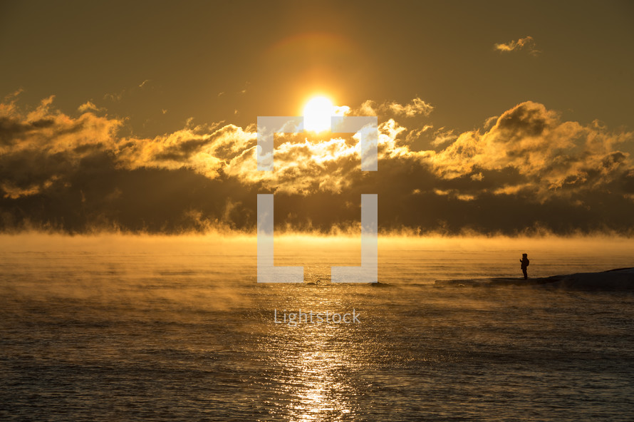 rising steam over ocean water and a man standing on a shore at sunrise 