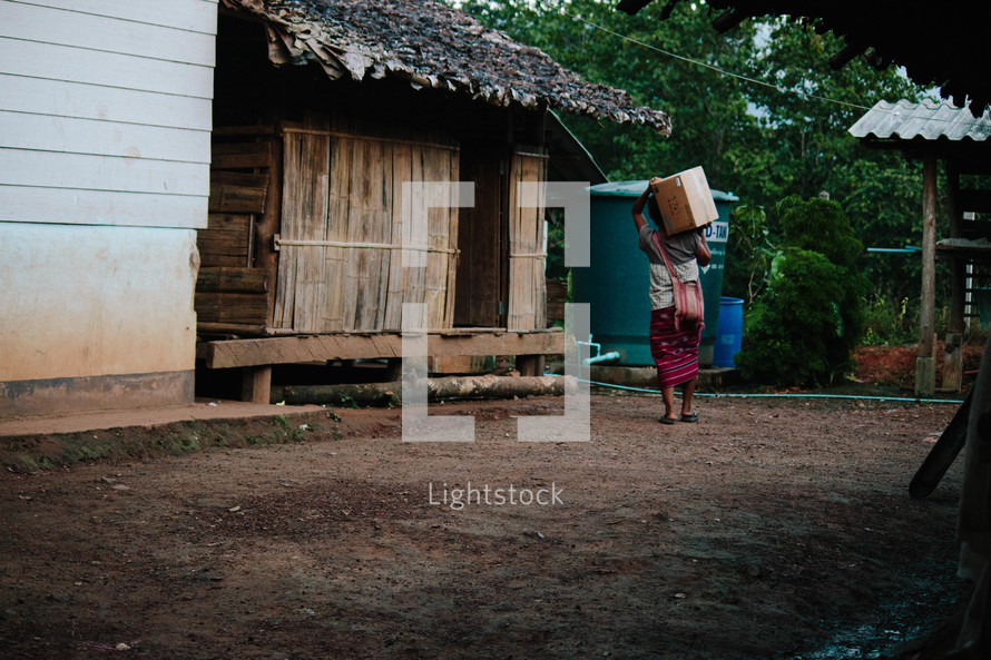 a woman walking carrying a box on a dirt road in a village 