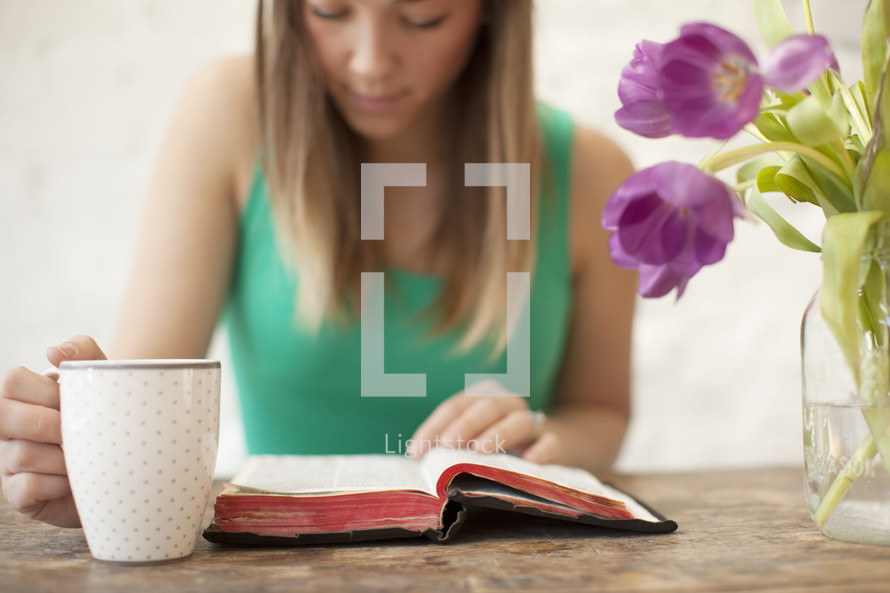 Woman with a coffee cup  reading the Bible on a wood table with tulips.