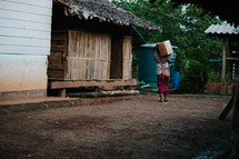 a woman walking carrying a box on a dirt road in a village 
