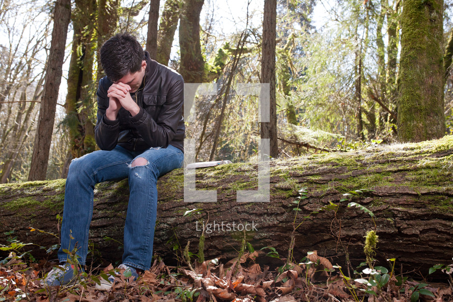 Man in prayer sitting on fallen tree in the woods.