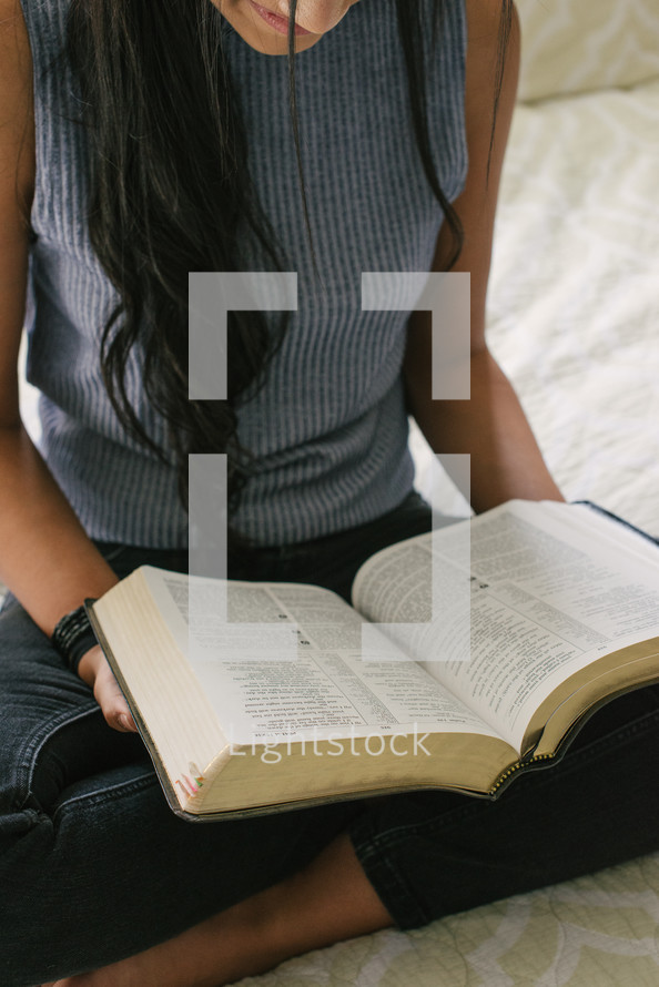 A young woman reading the Bible while sitting on her bed.