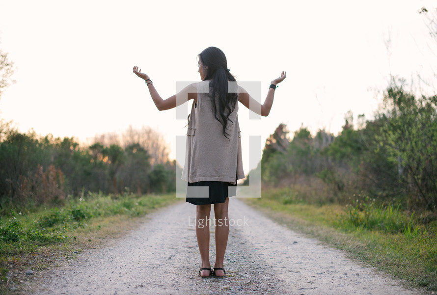 young woman standing on a gravel road with hands raised 