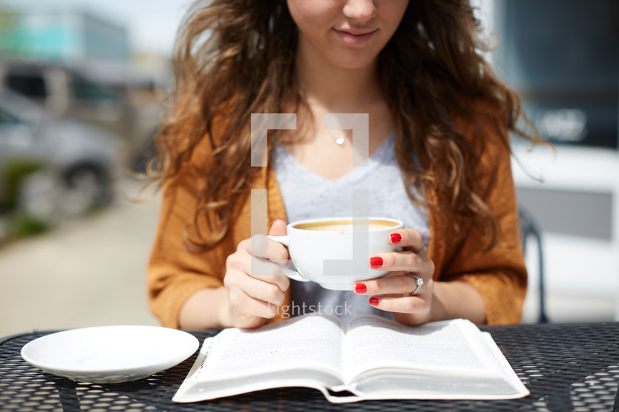 a woman sitting at an outdoor table reading a Bible and drinking coffee 