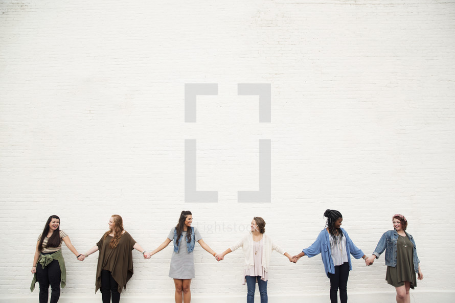 Six young women holding hands in a line in front of a white brick wall.