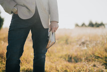 a man holding a Bible at his side outdoors in a field 