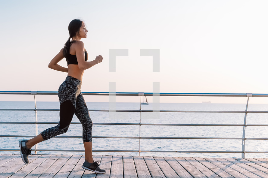 Athletic young girl jogging in morning by sea wooden embankment. Silhouette of girl in sports costume. Beautiful sunlight. Healthy lifestyle concept.