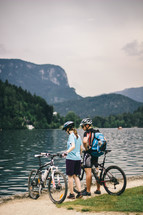 Two people with bicycles by a lake in the mountains.