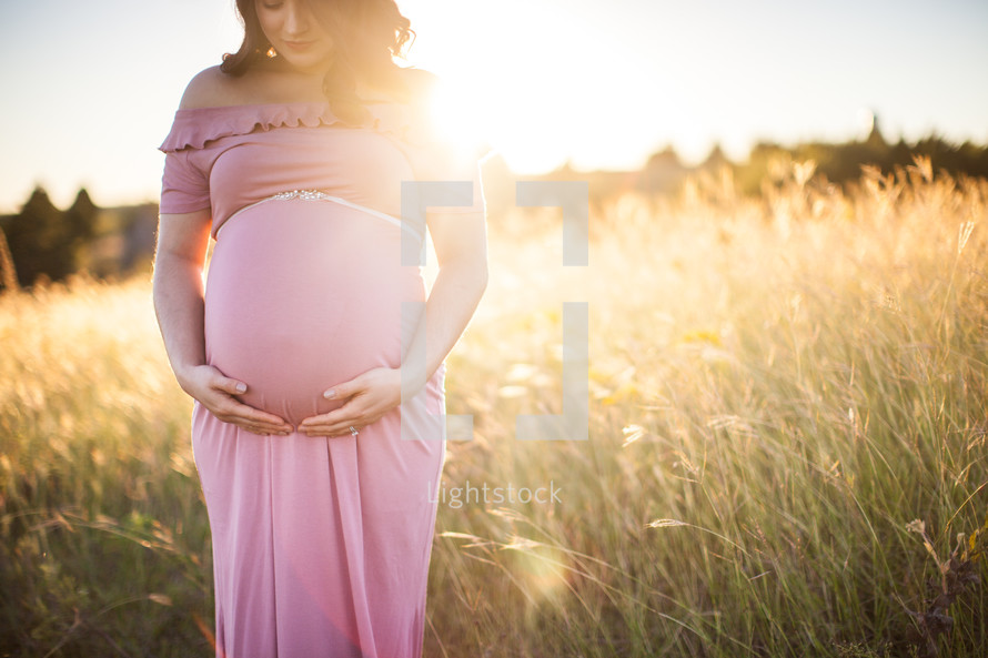 pregnant woman holding her belly standing in a field at sunset 