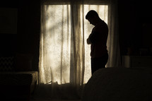 silhouette of a man standing in front of a window with head down thinking. 