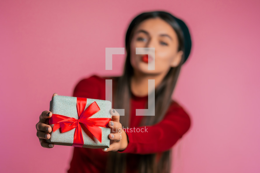 Pretty woman gives gift and hands it to the camera. She is happy, smiling. Girl on pink background. Positive holiday shot