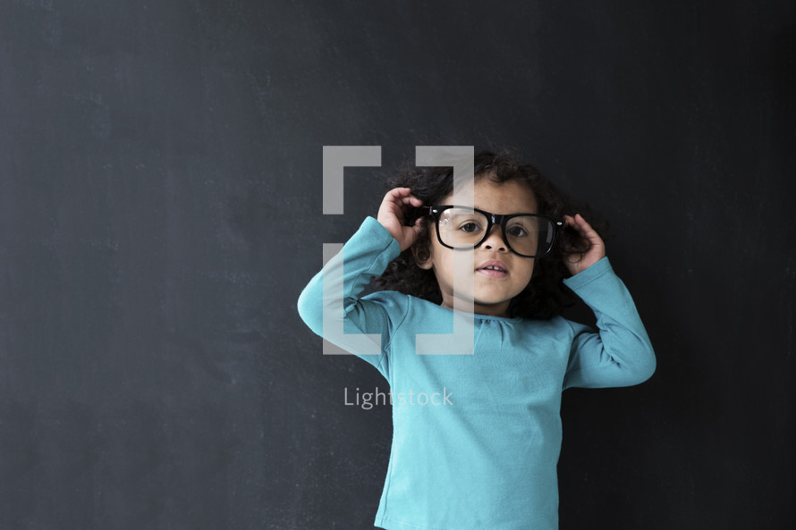 toddler girl wearing reading glasses against a chalkboard background.