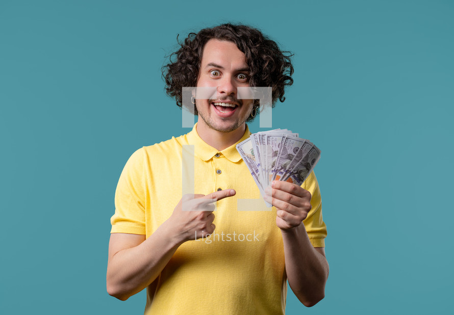 Satisfied man with USD currency. Young millenial american guy or student holding money - dollars banknotes on blue wall. Symbol of success, gain, salary, benefit, investments.