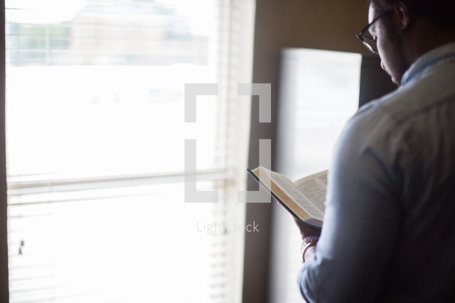 An African American man standing at a window reading a Bible 