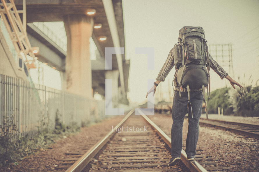 man balancing walking on railroad tracks with a backpack 