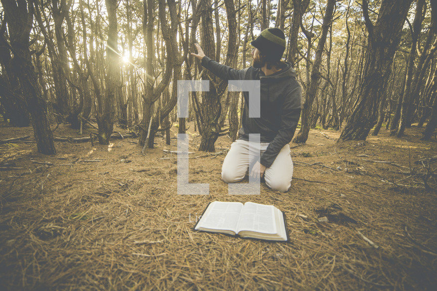 a man kneeling next to an open Bible on the ground in a forest 