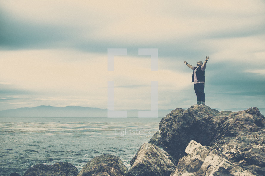 man standing on sea rocks with his hands raised in worship to God