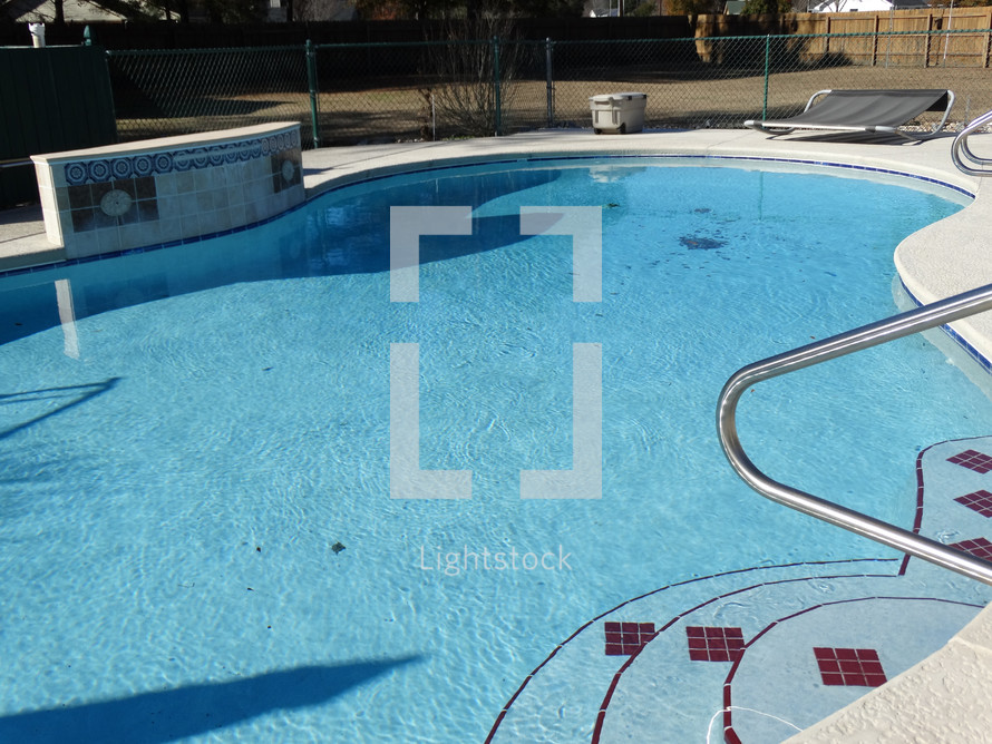 Swimming pool with bright blue water shining in sunlight surrounded by pool deck and waterfall wall