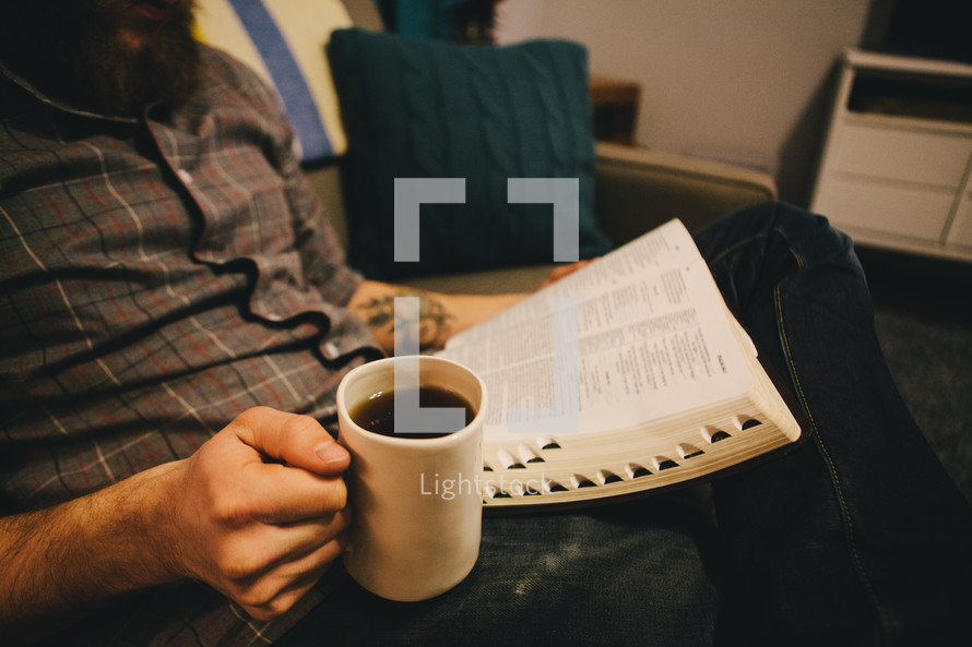 A man reading a Bible and holding a cup of coffee in his living room. 
