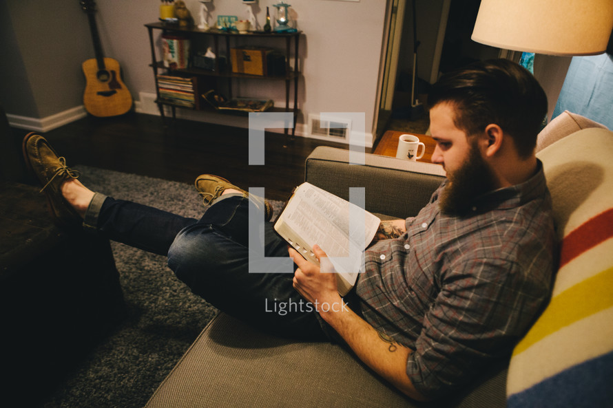 A man sitting on a couch in his living room reading a Bible. 