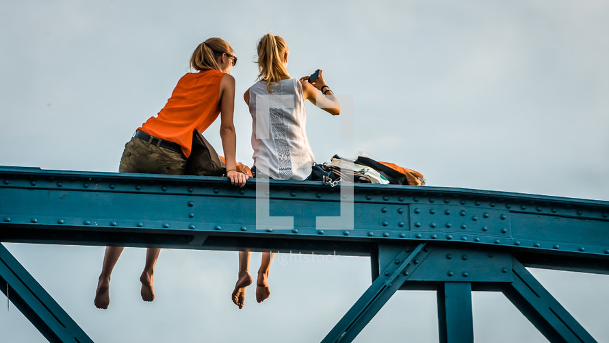 two women with feet dangling sitting on a bridge 