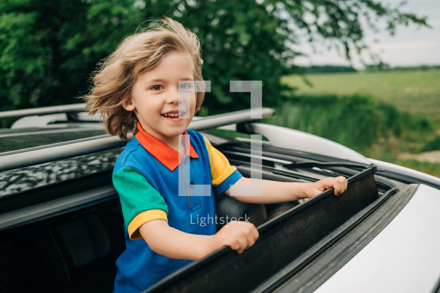 Adorable happy little boy stands in open car sunroof during road trip in countryside at summer. Concept of family leisure, active traveling. High quality photo