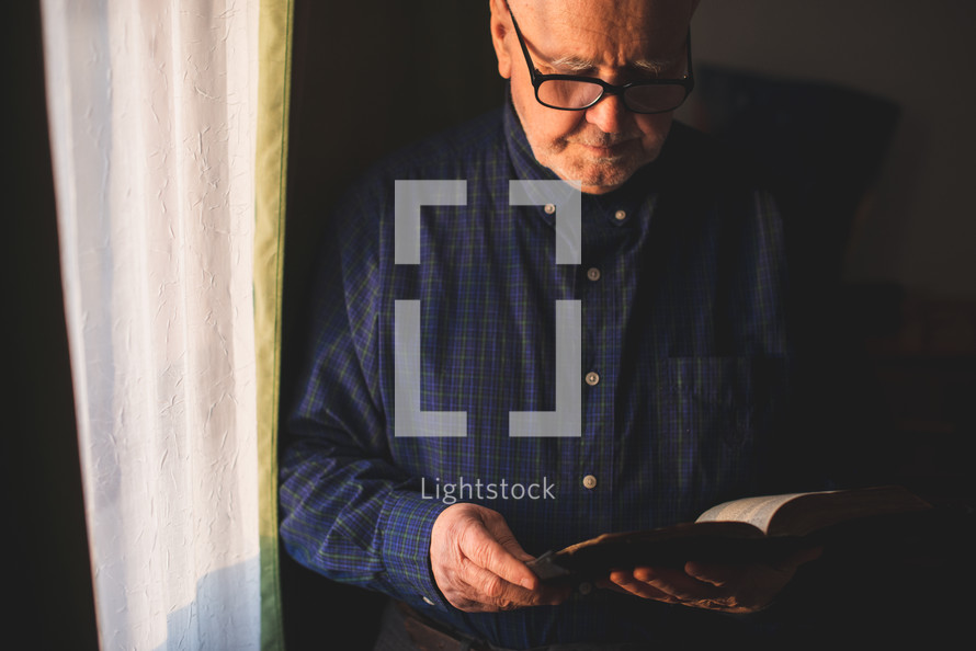 an elderly man reading a Bible standing by a window 