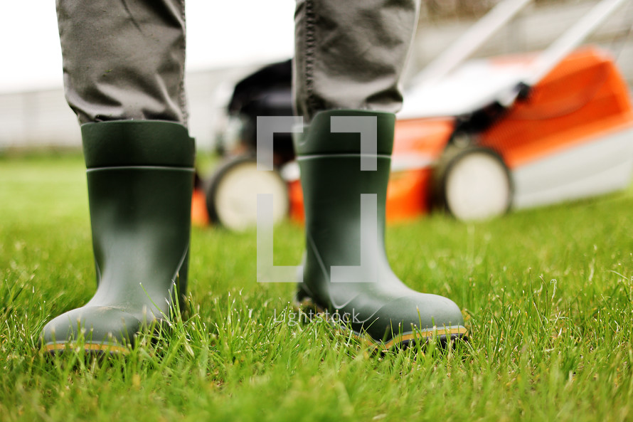 Cropped photo of male gardener legs in rubber boots stands on cut green grass lawn at backyard of house. Modern electric cordless lawnmower on back. Landscaping industry theme