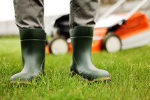 Cropped photo of male gardener legs in rubber boots stands on cut green grass lawn at backyard of house. Modern electric cordless lawnmower on back. Landscaping industry theme