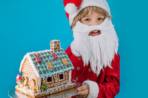 Cute little child in Santa Claus costume sitting with gingerbread house on blue background. Christmas celebration. Happy childhood, kid, lovely son. High quality photo