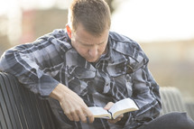 a man in a plaid shirt sitting on a bench outdoors reading a Bible 