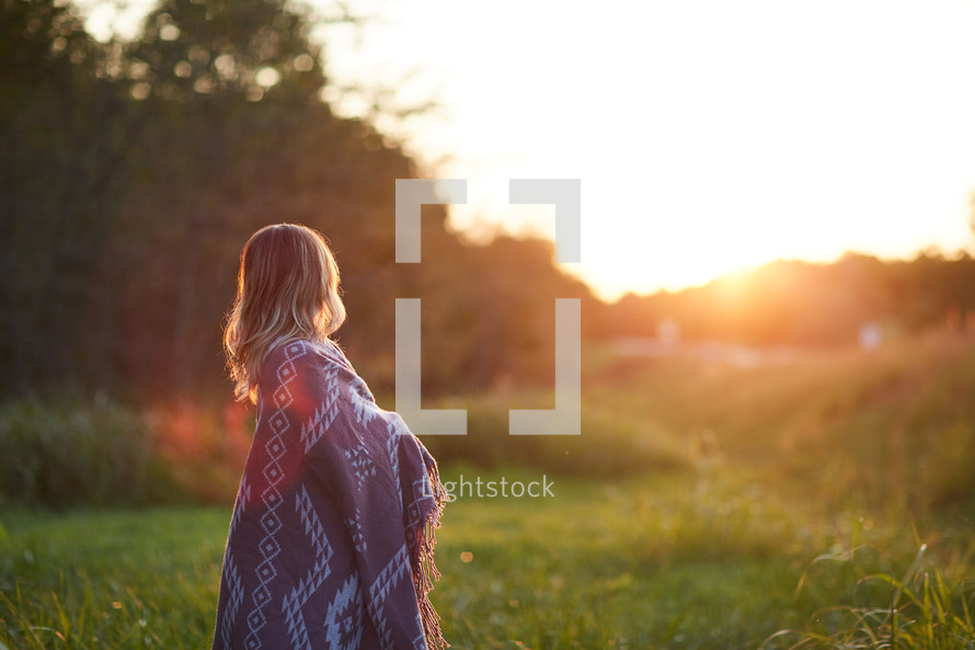a woman wrapped in a blanket standing outdoors at sunset 