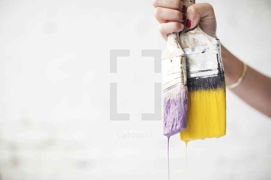 A woman holds two dripping paint brushes.