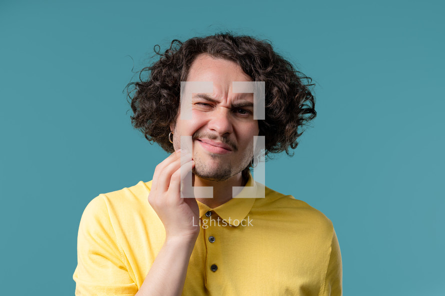 Handsome curly haired man with tooth pain on blue studio background. Toothache, dental problems, dentistry and medicine concept