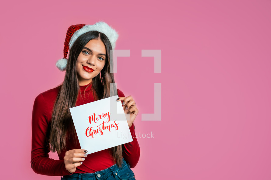 Attractive girl in Santa hat with nameplate Merry Christmas. Portrait of lady on pink background. New year concept. Happy pretty woman smiling to camera