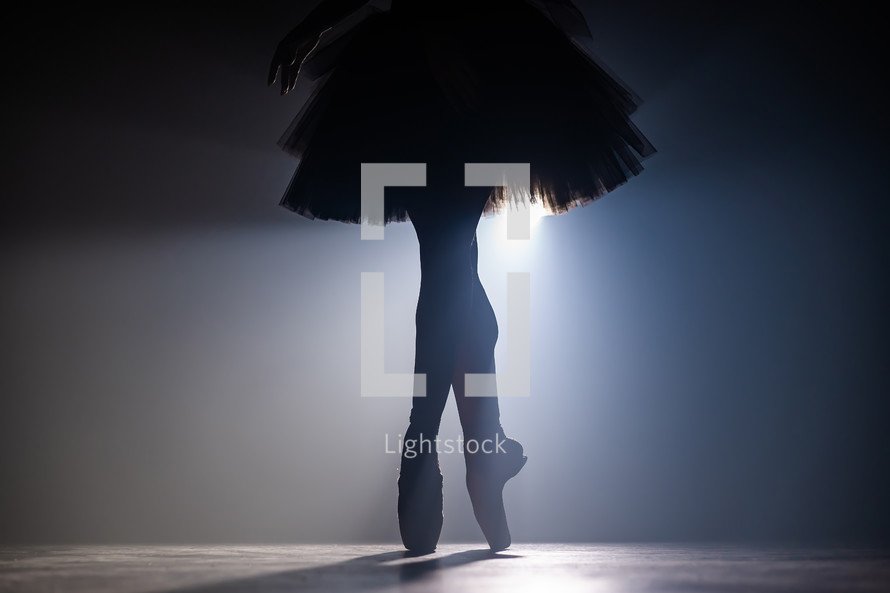 Close up silhouette of ballerina legs in tutu dress. Ballet performance on dark stage with floodlight backlight. Smoke background