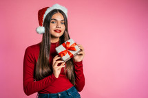 Attractive girl in Santa hat with gift box. Portrait of lady on pink background. Christmas or New year concept. Happy pretty woman smiling to camera