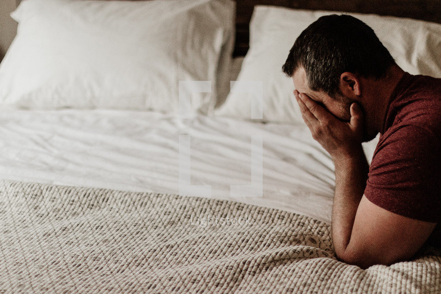 a man covering his face kneeling in prayer over a bed 