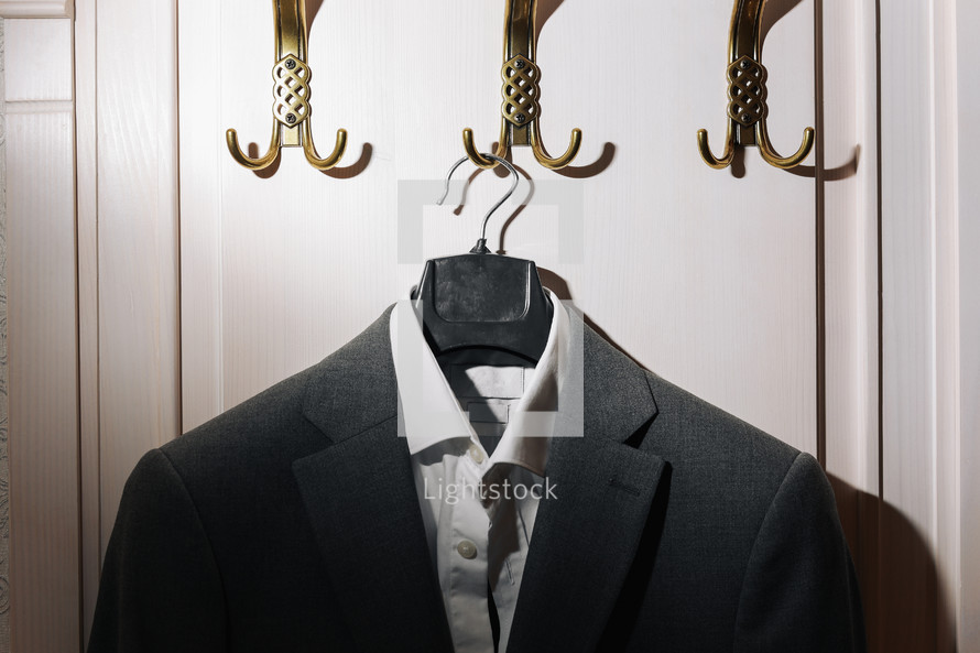 Man's dark grey suit jacket and white shirt hanging on gold metal hook on white wall background