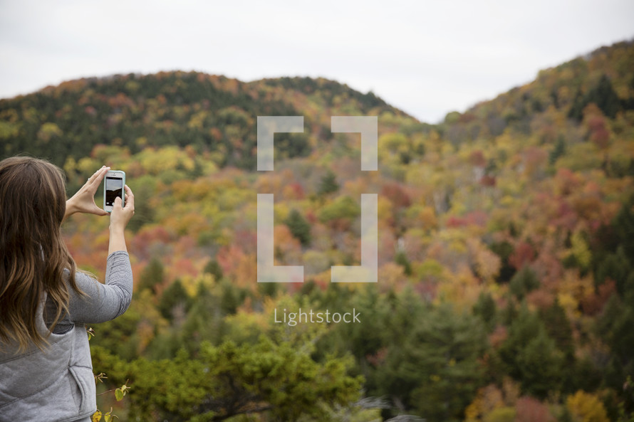woman taking a picture with her phone of a fall mountain forest 