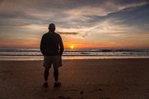 A man standing on a beach and watching the sun set over the ocean.
